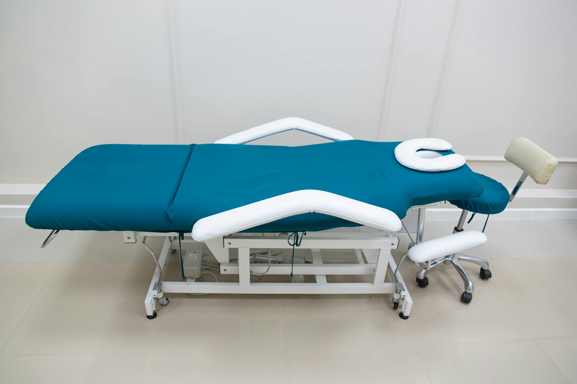 Wide angle view of a professional chiropractic clinic room, adjusting table centered, neutral walls with calming teal accents, clean modern medical equipment, soft natural light from window, clinical but warm and inviting, photorealistic interior photography