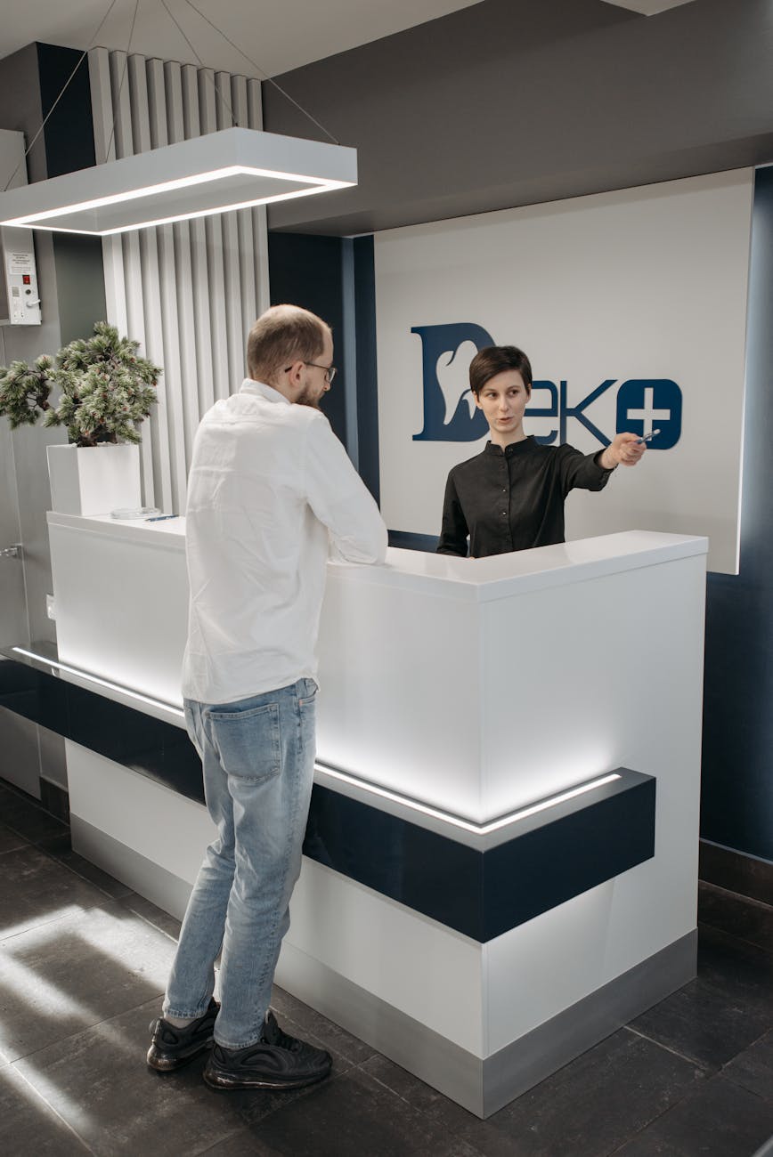 Friendly medical receptionist greeting a new patient at a clean modern clinic front desk, warm welcoming atmosphere, teal and white interior, natural lighting, candid professional photograph, reassuring and approachable scene, photorealistic