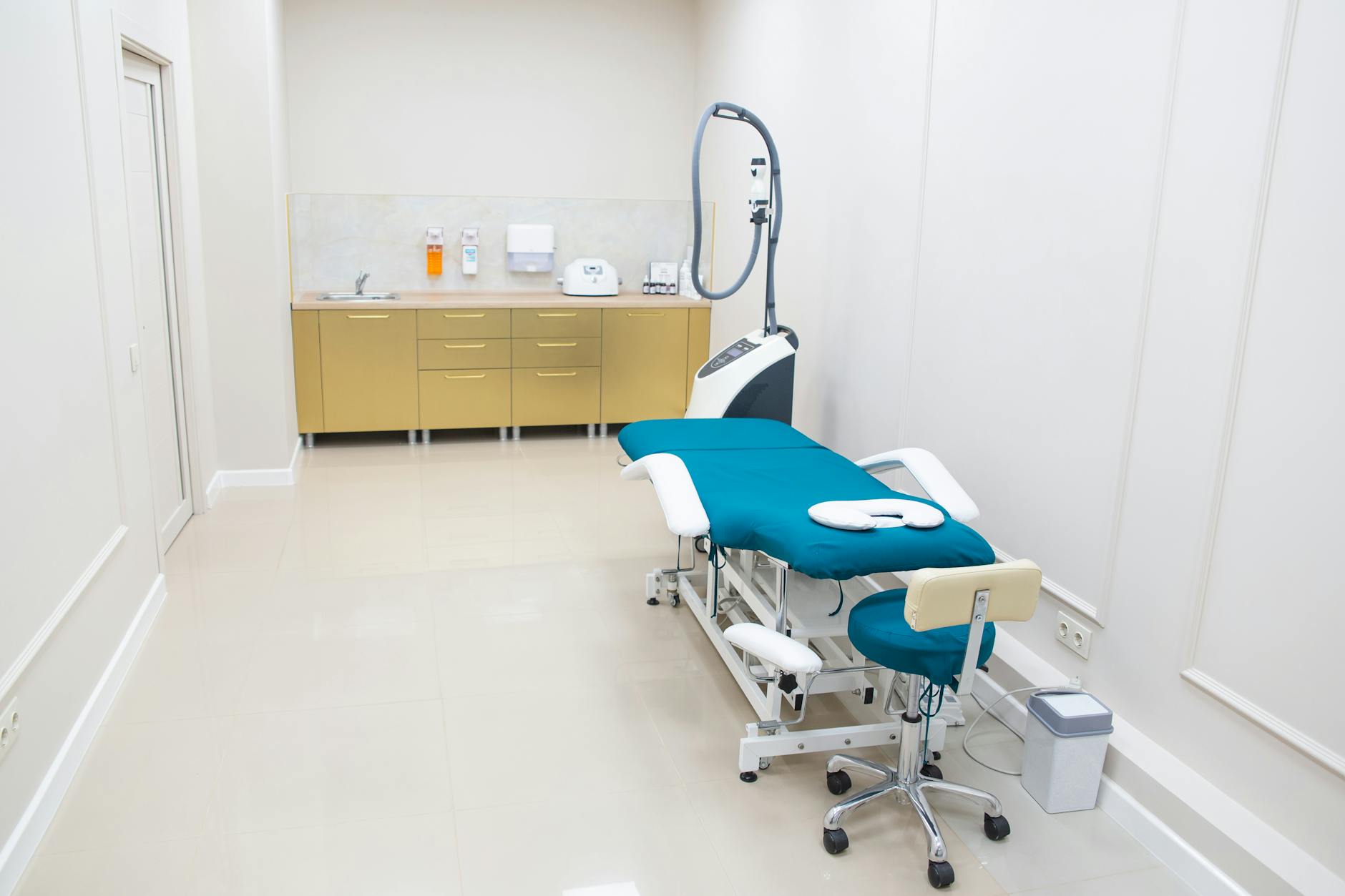 Podiatry clinic treatment room with specialized examination chair, foot care instruments neatly arranged, bright clean white and teal interior, professional medical setting, warm lighting, modern podiatric office, photorealistic interior photography