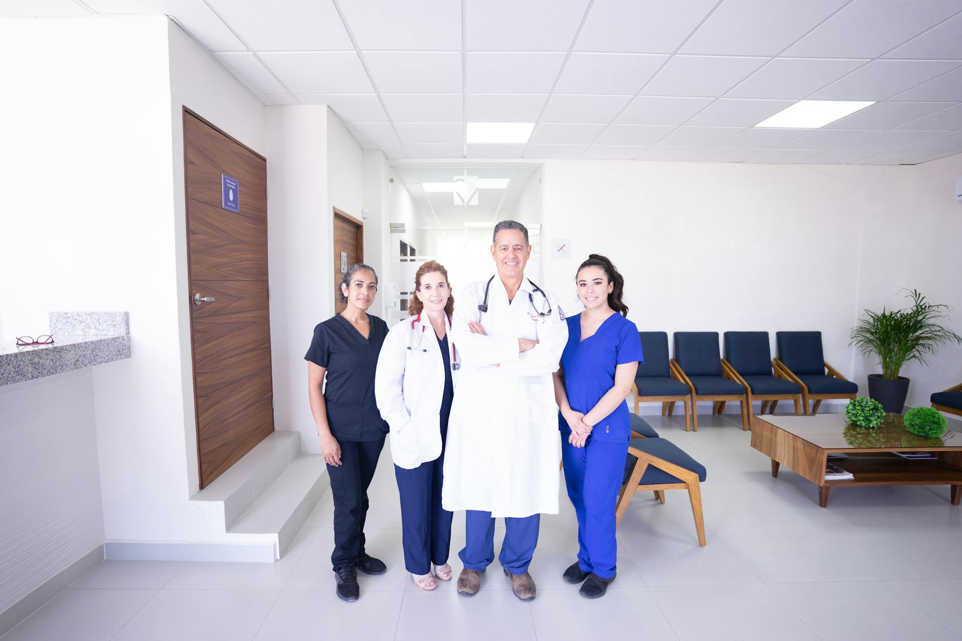 Three medical professionals in white coats standing together in a bright modern medical office lobby, warm smiles, professional and approachable, Long Beach California suburban clinic, warm interior lighting, photorealistic group portrait